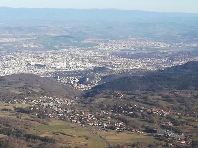 A high-angle panoramic photograph looking down from a mountain summit onto a sprawling urban area (Clermont-Ferrand) and rolling countryside, with distant mountain ranges on the horizon.