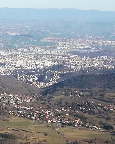 A high-angle panoramic photograph looking down from a mountain summit onto a sprawling urban area (Clermont-Ferrand) and rolling countryside, with distant mountain ranges on the horizon.