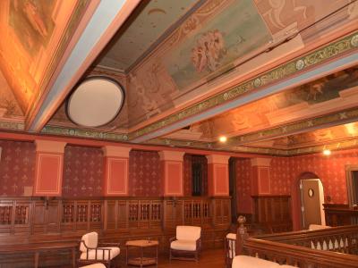 The image shows an ornate interior room with painted ceilings, red patterned wallpaper, wooden wainscoting, and white upholstered chairs, likely a historic hall or museum space.