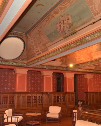 The image shows an ornate interior room with painted ceilings, red patterned wallpaper, wooden wainscoting, and white upholstered chairs, likely a historic hall or museum space.