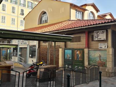 A street-level view of a building with a terracotta-tiled roof and stone base, featuring a green awning for "Le temps des oliviers" and signage for "Les Halles" and "Des halles," with a scooter parked in front.