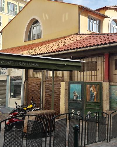 A street-level view of a building with a terracotta-tiled roof and stone base, featuring a green awning for "Le temps des oliviers" and signage for "Les Halles" and "Des halles," with a scooter parked in front.