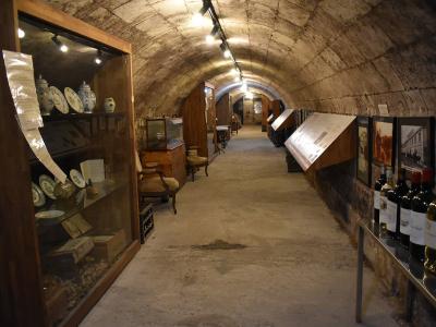A long, arched stone cellar or tunnel repurposed as a museum exhibit, featuring display cases with antique ceramics and furniture on the left, and framed historical photos alongside rows of wine bottles on the right.