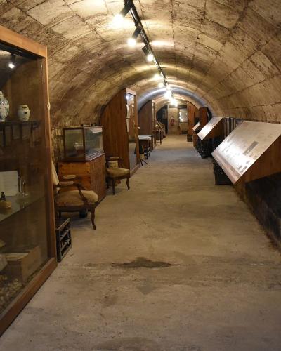 A long, arched stone cellar or tunnel repurposed as a museum exhibit, featuring display cases with antique ceramics and furniture on the left, and framed historical photos alongside rows of wine bottles on the right.