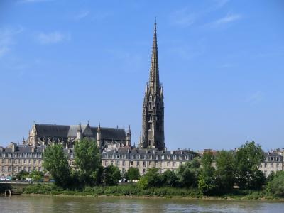 A Gothic church with a tall, ornate spire and flying buttresses, viewed from across a river with trees and classical buildings in the foreground under a blue sky.