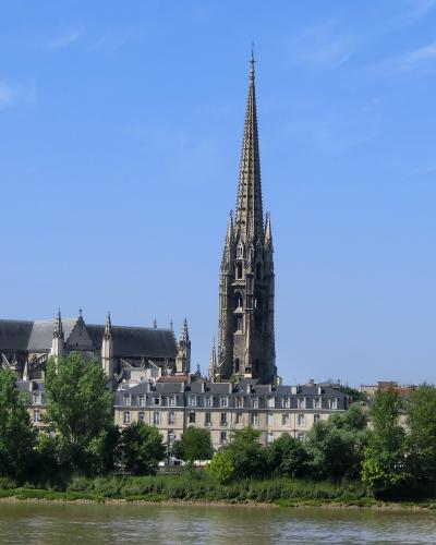 A Gothic church with a tall, ornate spire and flying buttresses, viewed from across a river with trees and classical buildings in the foreground under a blue sky.