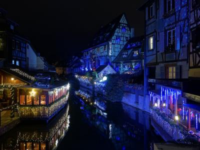 A night view of a canal lined with half-timbered buildings adorned with festive lights, featuring a prominent vertical "HOTEL" sign and a building labeled "LES BATTEURS" on the left bank.