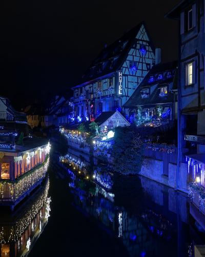 A night view of a canal lined with half-timbered buildings adorned with festive lights, featuring a prominent vertical "HOTEL" sign and a building labeled "LES BATTEURS" on the left bank.