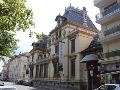The image shows the Château Lumière, a grand late 19th-century mansion with distinctive architecture including a steeply pitched roof and ornate details, with vertical banners hanging from the facade that read "Institut Lumière" and feature images