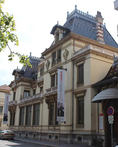 The image shows the Château Lumière, a grand late 19th-century mansion with distinctive architecture including a steeply pitched roof and ornate details, with vertical banners hanging from the facade that read "Institut Lumière" and feature images