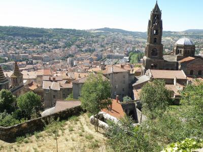 A panoramic view of a historic French city featuring a prominent stone cathedral with a tall bell tower and a dome, overlooking a dense urban area with terracotta roofs and rolling green hills in the background.