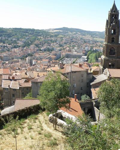A panoramic view of a historic French city featuring a prominent stone cathedral with a tall bell tower and a dome, overlooking a dense urban area with terracotta roofs and rolling green hills in the background.