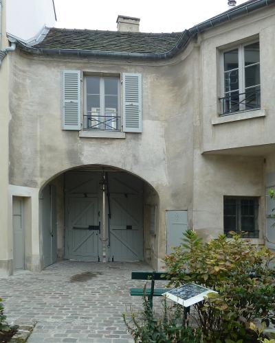 A garden scene with a manicured lawn, a distinctive low-slung tree supported by a metal stand, a stone wall, a cobblestone path, and a green lamppost, with buildings in the background.