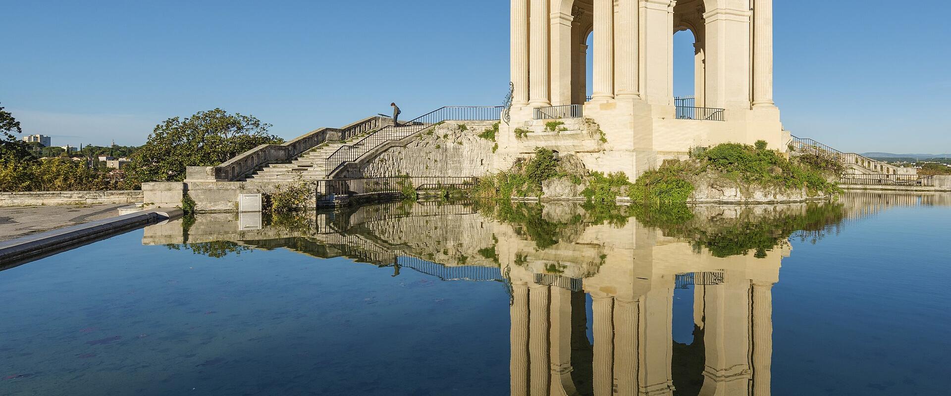 1. The image shows the Château d'eau du Peyrou, a classical stone pavilion with columns and arches, situated on a raised platform beside a reflecting pool in Montpellier, France, under a clear blue sky.