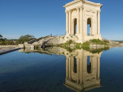 1. The image shows the Château d'eau du Peyrou, a classical stone pavilion with columns and arches, situated on a raised platform beside a reflecting pool in Montpellier, France, under a clear blue sky.