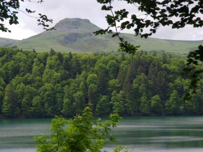 A calm lake in the foreground with a dense green forest along the far shore and a prominent, rounded volcanic cone rising behind the trees under an overcast sky.