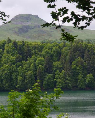 A calm lake in the foreground with a dense green forest along the far shore and a prominent, rounded volcanic cone rising behind the trees under an overcast sky.