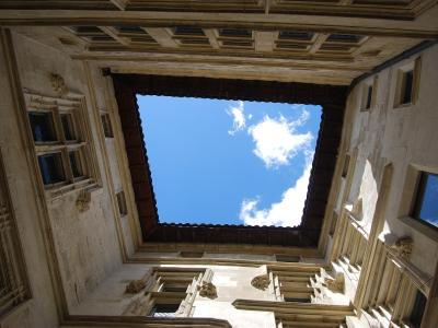 A low-angle architectural photograph looking up into a stone courtyard or atrium of a historic building, featuring Renaissance-style windows, carved stone gargoyles or grotesques, and a dark wooden roof structure framing a blue sky with clouds.