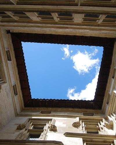 A low-angle architectural photograph looking up into a stone courtyard or atrium of a historic building, featuring Renaissance-style windows, carved stone gargoyles or grotesques, and a dark wooden roof structure framing a blue sky with clouds.