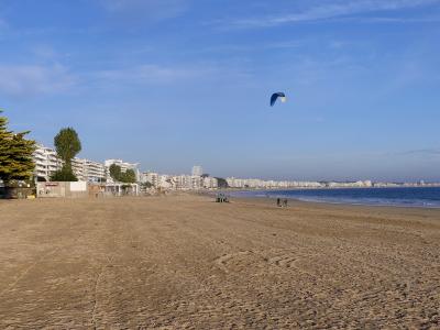 Wide sandy beach with a blue kite surf sail and coastal buildings under a blue sky.