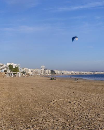 Wide sandy beach with a blue kite surf sail and coastal buildings under a blue sky.