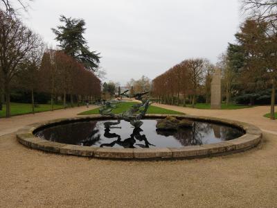 A circular stone-edged pond with a dark, abstract metal sculpture in the center, set within a gravel path and flanked by rows of trees and a tall stone obelisk on the right.