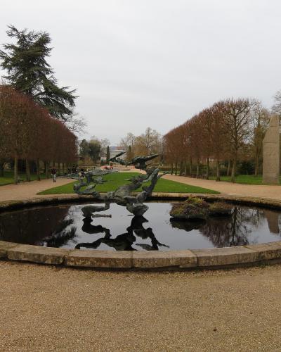 A circular stone-edged pond with a dark, abstract metal sculpture in the center, set within a gravel path and flanked by rows of trees and a tall stone obelisk on the right.