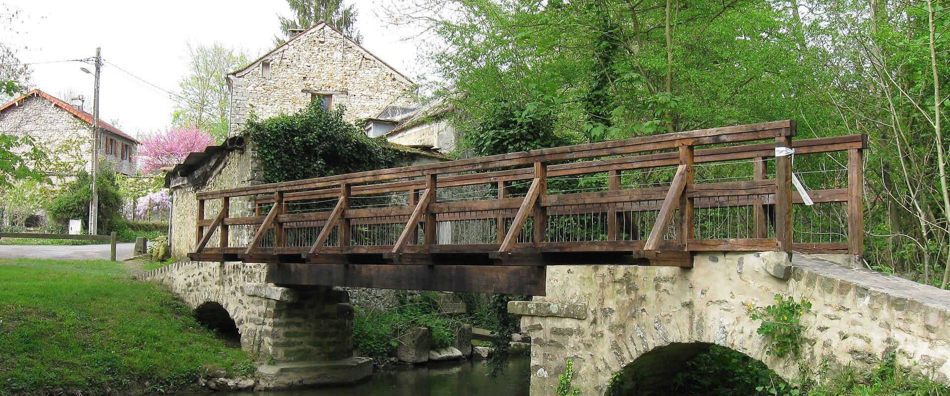 The image shows a stone bridge with a wooden pedestrian walkway and railings spanning a small, calm river, flanked by green grass and trees, with a stone building visible in the background on the left bank.