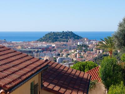 A high-angle view of the Nice coastline featuring a prominent green hill (Castle Hill) overlooking the Mediterranean Sea, with terracotta rooftops in the foreground and the filename indicating the photo was taken from Cimiez.