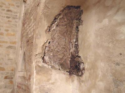 The image shows an interior architectural detail of a stone wall featuring a recessed niche containing a fragment of ancient, faded geometric or vine-like fresco painting, with exposed brickwork visible on the left side.