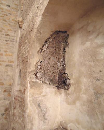 The image shows an interior architectural detail of a stone wall featuring a recessed niche containing a fragment of ancient, faded geometric or vine-like fresco painting, with exposed brickwork visible on the left side.