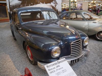 A dark blue 1953 Hotchkiss-Grégoire Berline JAG on display in a museum setting, with an informational placard identifying the car and its origin (France), and another vintage car visible in the background.