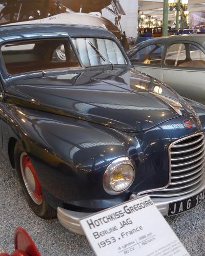 A dark blue 1953 Hotchkiss-Grégoire Berline JAG on display in a museum setting, with an informational placard identifying the car and its origin (France), and another vintage car visible in the background.