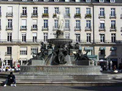 A large, ornate bronze fountain with multiple sculpted figures and a central statue on top, situated in a paved public square in front of a multi-story neoclassical building with uniform windows and balconies; people are seated at outdoor cafes ne