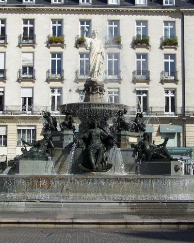 A large, ornate bronze fountain with multiple sculpted figures and a central statue on top, situated in a paved public square in front of a multi-story neoclassical building with uniform windows and balconies; people are seated at outdoor cafes ne