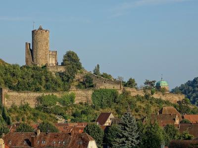 A stone castle ruin with a prominent round tower sits atop a forested hill overlooking a town with red-tiled roofs and a green-domed church in the background.