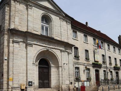 A low-angle photograph of a large, light-colored stone building with a classical arched entrance, pediment, and a long wing extending to the right, featuring French flags and flower boxes.