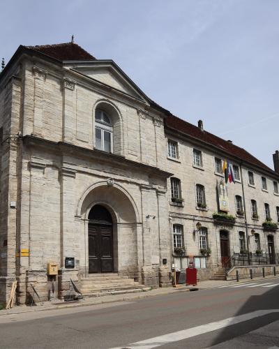 A low-angle photograph of a large, light-colored stone building with a classical arched entrance, pediment, and a long wing extending to the right, featuring French flags and flower boxes.