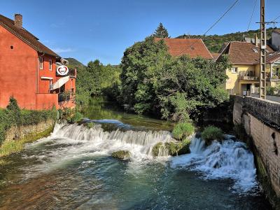 A scenic view of a river with a small weir or cascade flowing through a town, featuring a prominent red building on the left bank and stone embankments, with a sign for "La Cave des Vignerons" visible on the red building.