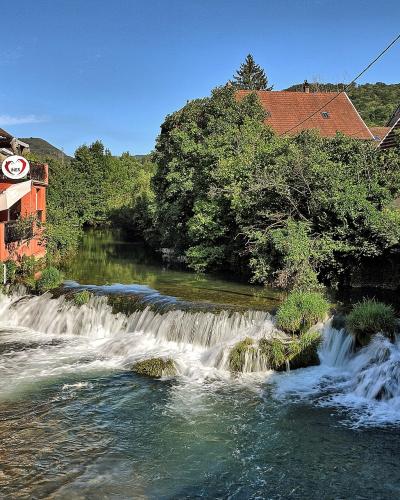 A scenic view of a river with a small weir or cascade flowing through a town, featuring a prominent red building on the left bank and stone embankments, with a sign for "La Cave des Vignerons" visible on the red building.
