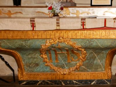 The image shows an ornate church altar with a green marble front panel featuring a golden IHS monogram wreath, draped in embroidered liturgical cloth, with candles and flowers on the mensa.
