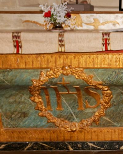 The image shows an ornate church altar with a green marble front panel featuring a golden IHS monogram wreath, draped in embroidered liturgical cloth, with candles and flowers on the mensa.