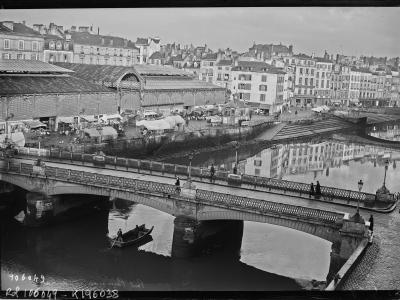 A black and white historical photograph showing a stone bridge spanning a river with a rowboat underneath, and a large, long covered market hall with an arched roof structure situated on the riverbank behind the bridge.