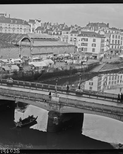 A black and white historical photograph showing a stone bridge spanning a river with a rowboat underneath, and a large, long covered market hall with an arched roof structure situated on the riverbank behind the bridge.
