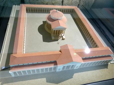 A detailed architectural scale model of a Gallo-Roman temple complex with a central circular temple surrounded by a colonnaded courtyard and adjacent buildings, displayed in a glass case.