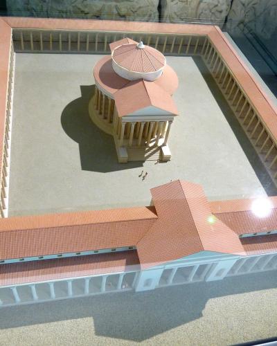 A detailed architectural scale model of a Gallo-Roman temple complex with a central circular temple surrounded by a colonnaded courtyard and adjacent buildings, displayed in a glass case.