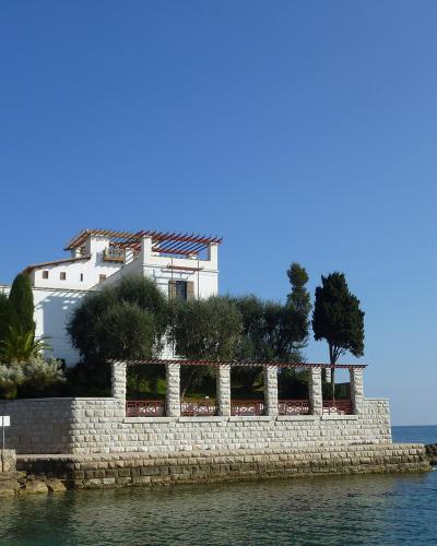 A white, multi-story villa with a pergola and stone retaining wall situated on the coast, featuring a colonnaded terrace structure in the foreground.