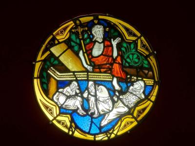 A circular stained-glass medallion depicting the Resurrection of Jesus, showing Christ rising from a tomb while three sleeping guards lie below, set against a dark background.