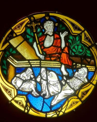 A circular stained-glass medallion depicting the Resurrection of Jesus, showing Christ rising from a tomb while three sleeping guards lie below, set against a dark background.