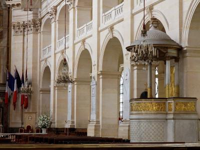 The interior of a grand stone church featuring a prominent marble and gold pulpit, arched colonnades, hanging chandeliers, and French flags.